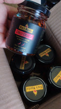 Load image into Gallery viewer, Black Mamba Chilli Crunch, with several other jars stacked inside a cardboard box in the background.