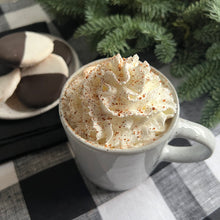 Load image into Gallery viewer, White mug with whipped cream and ancho habanero powder on a checked tablecloth with buscuits and a plant in the background.