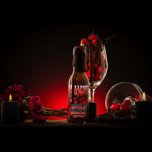 Load image into Gallery viewer, Chocolate Cherry Sauce stands on a dark tabletop beside a large wine glass filled with red cherries, candles, and red roses against a black background with red lighting.
