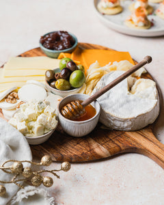 Wooden platter with assorted cheeses, olives, and hot honey on a light background