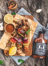 Load image into Gallery viewer, Skewers of grilled meat with lemon wedges, a bottle of Hellfire Elixir sauce, and a small bowl of sauce on a wooden surface.
