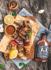 Skewers of grilled meat with lemon wedges, a bottle of Hellfire Elixir sauce, and a small bowl of sauce on a wooden surface.