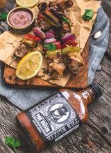 Load image into Gallery viewer, Wooden cutting board with skewers of grilled meat and vegetables, accompanied by a bottle of Hellfire Elixir hot sauce
